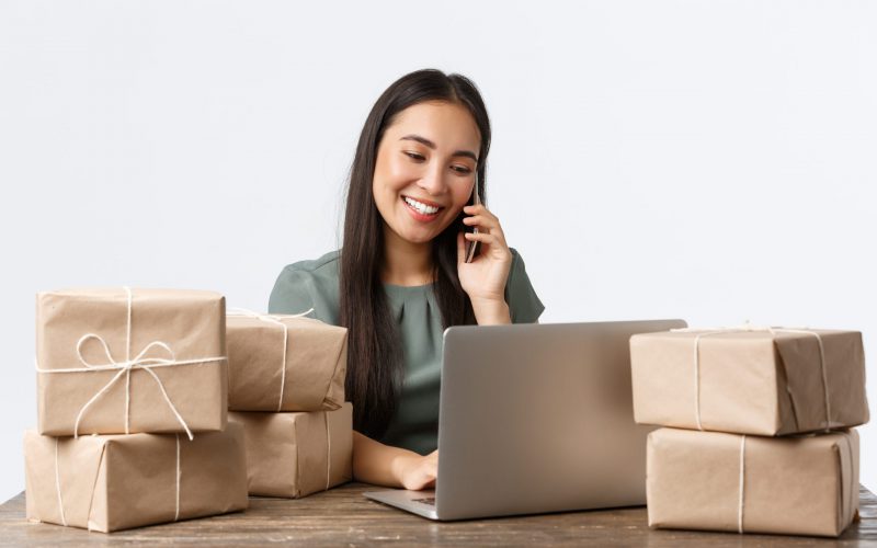 Small business owners, startup and e-commerce concept. Smiling asian female store manager, entrepreneur with online shop answering client calls, managing orders via laptop, packing items for shipping.